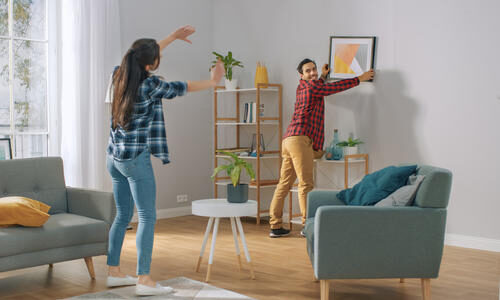 happy-couple-hanging-picture-on-the-wall-boyfriend-moves-it-girlfriend-tells-him-when-the-frame-is-hanging-straight-funny-moment-in-young-couples-life-modern-stylish-apartment